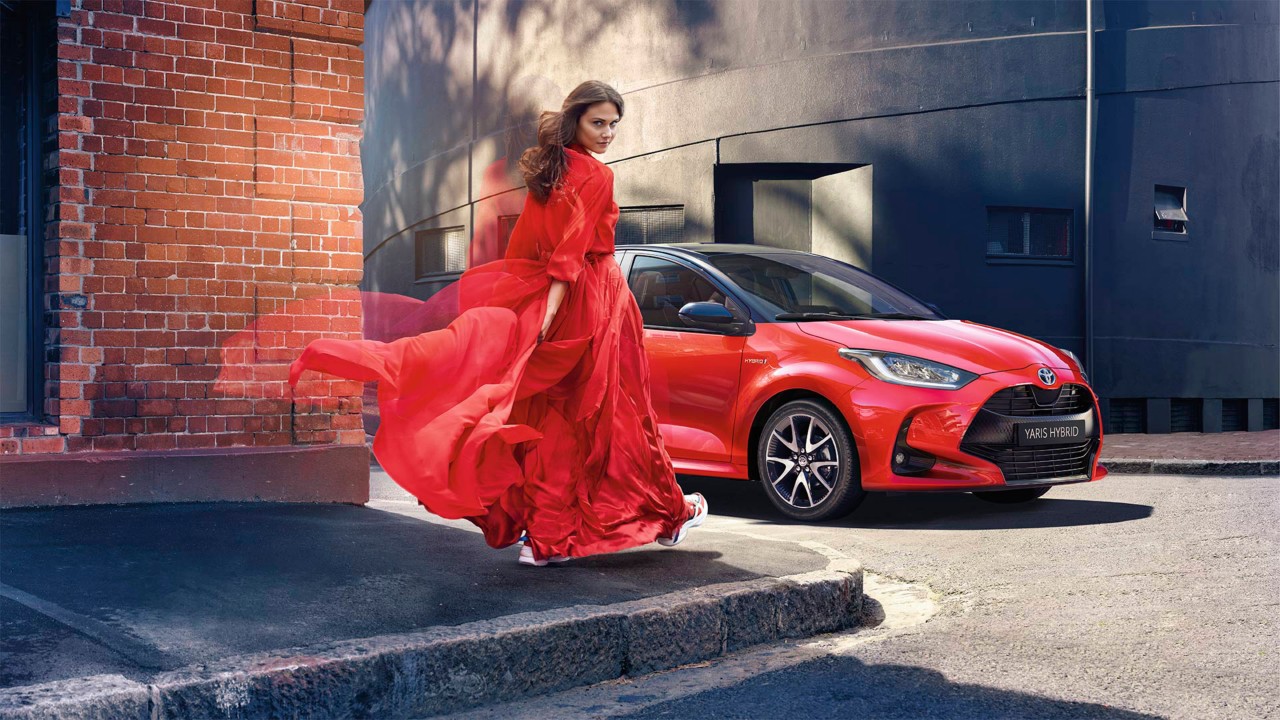 En kvinna i röd klänning bredvid en röd Toyota Yaris bil.