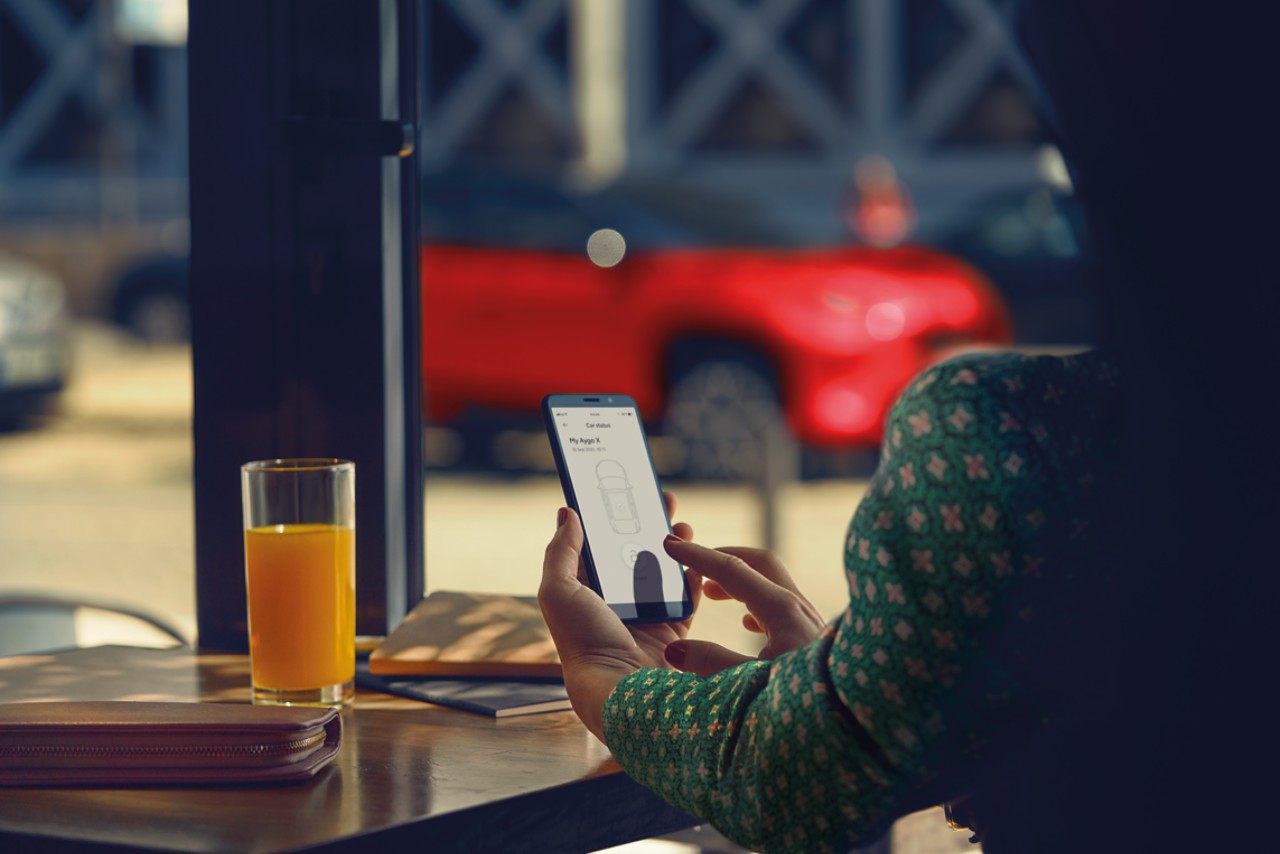 En kvinna sitter på ett café och använder MyToyota-appen på sin mobiltelefon. I bakgrunden syns hennes röda Toyota Yaris Cross.