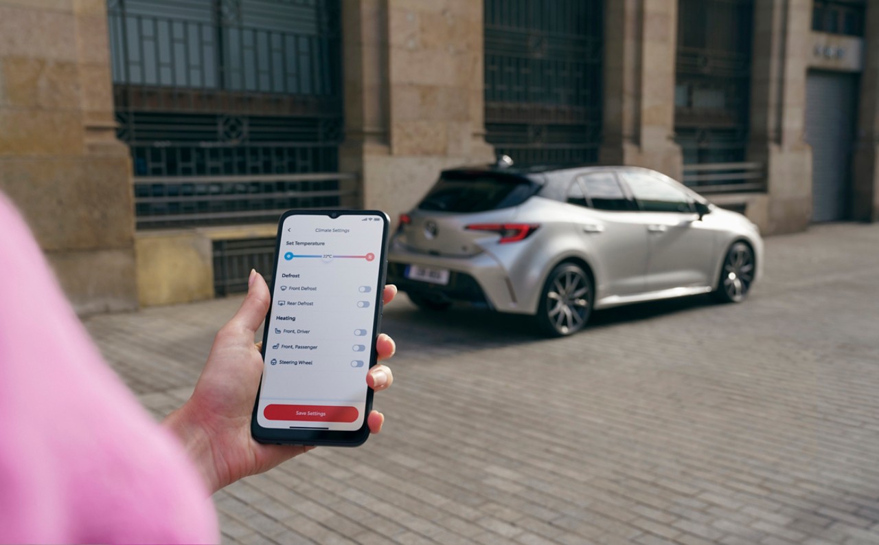 En person håller i sin mobiltelefon som visar MyToyota-appen. I bakgrunden står en Toyota Corolla parkerad.